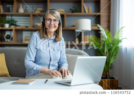 Smiling senior woman wearing headphones with microphone, working remotely from home laptop. Confident professional communicating online, participating virtual meeting. Cozy home office environment. 116543752