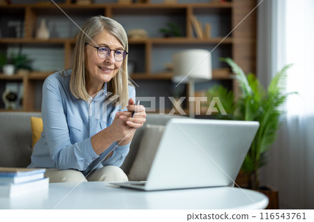 Elderly woman sitting on sofa having online video call using laptop. Mature lady wearing glasses smiling while chatting, engaging in digital communication from cozy indoor space. 116543761