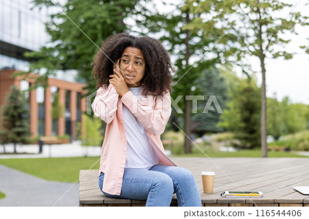 Young woman student experiencing ear pain while sitting outdoors. She holds her ear with a pained expression, suggesting discomfort from otitis or ear inflammation. Young woman student experiencing ear pain while sitting outdoors. She holds her ear with a pained expression, suggesting discomfort from otitis or ear inflammation. 116544406
