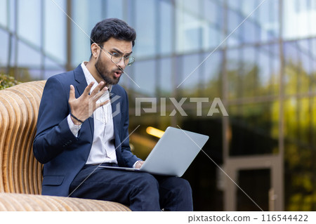 Hispanic businessman wearing suit surprised during work on laptop outdoors. Emotionally engaged in online communication or study. Urban setting with modern building in background. Hispanic businessman wearing suit surprised during work on laptop outdoors. Emotionally engaged in online communication or study. Urban setting with modern building in background. 116544422