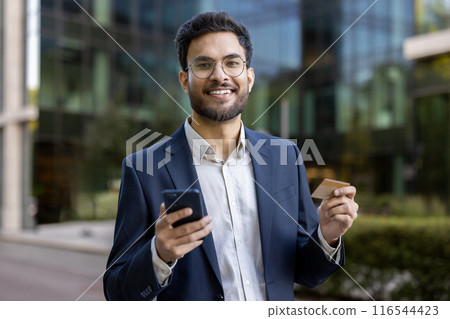 Hispanic businessman in suit standing outside modern office building. Man holding smartphone and credit card, smiling confidently. Concept of digital transactions, business, finance and technology. 116544423