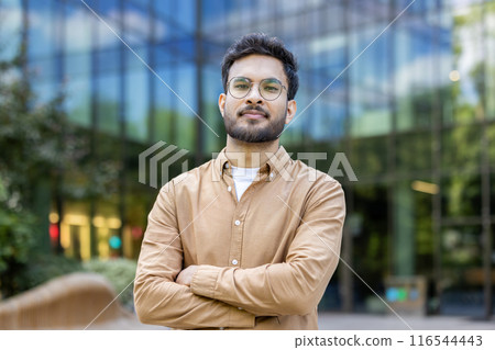 Confident hispanic man with glasses standing outside modern glass building with arms crossed, looking at camera with expression assurance and professionalism. Concept business, success, confidence 116544443