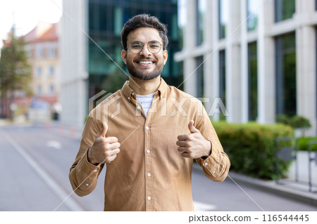 Smiling hispanic man wearing casual outfit giving thumbs up on city street. Young professional showing positivity, confidence, and approval outdoors urban environment. Modern buildings background. 116544445
