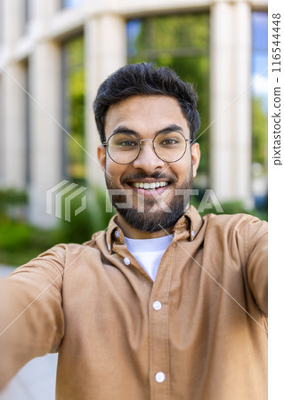 Cheerful hispanic man with glasses taking selfie outside modern office building on sunny day, smiling at camera. Business casual attire, outdoor setting with green background. 116544448