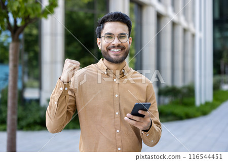 Confident hispanic man wearing glasses holding smartphone outdoors in urban setting, celebrating success with fist pump. Professional and positive emotion in business environment. Confident hispanic man wearing glasses holding smartphone outdoors in urban setting, celebrating success with fist pump. Professional and positive emotion in business environment. 116544451