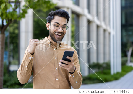 Hispanic man feeling excited and celebrating success while using smartphone outside in city area. Casual guy with glasses smiling and showing fist in triumph. Hispanic man feeling excited and celebrating success while using smartphone outside in city area. Casual guy with glasses smiling and showing fist in triumph. 116544452