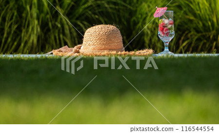 Straw hat and a glass with a refreshing drink by the pool, with green grass in the background 116544554