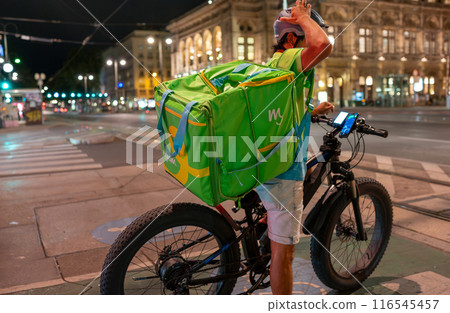 Vienna, Austria August 20, 2022. Night shot of a worker from the delivery rider company mjam. Riding his e-bike, he waits for the green light to enter traffic. In the background the Vienna Opera. Vienna, Austria August 20, 2022. Night shot of a worker from the delivery rider company mjam. Riding his e-bike, he waits for the green light to enter traffic. In the background the Vienna Opera. 116545457