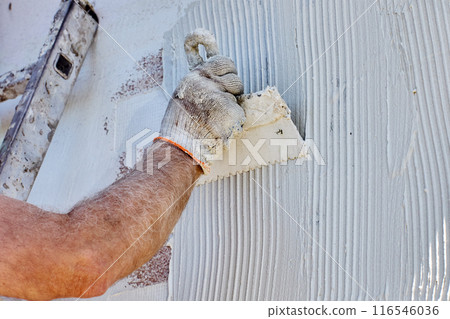 Applying tile adhesive to facade of building, house painter uses steel notched trowel. Applying tile adhesive to facade of building, house painter uses steel notched trowel. 116546036