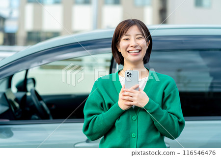 Smiling woman holding a smartphone in front of a car Smartphone and car Smiling woman holding a smartphone in front of a car Smartphone and car 116546476