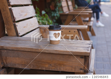 paper cup of natural coffee with carved white heart stands on armrest of wooden chair on city street 116546843