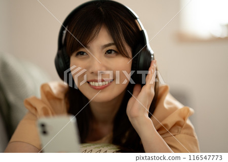 A middle-aged woman lying on a sofa listening to music on her smartphone 116547773