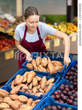 Smiling saleswoman lying out boxes of sweet potatoes in farmers market Smiling saleswoman lying out boxes of sweet potatoes in farmers market 116548808