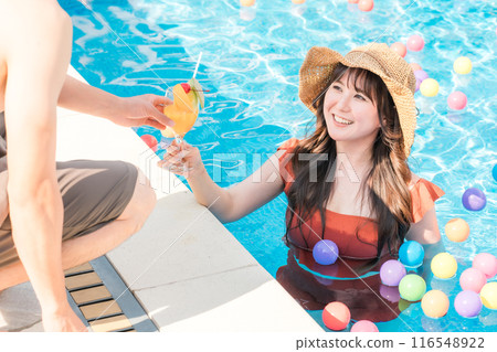 一位穿著泳衣的女士在游泳池裡度假,並從泳池邊的一位男士那裡收到了熱帶飲料。 一位穿著泳衣的女士在游泳池裡度假,並從泳池邊的一位男士那裡收到了熱帶飲料。 116548922