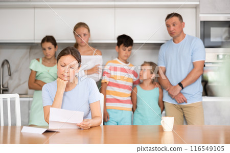 Upset mum is sitting in kitchen, reading letter. Blurred worried family in background 116549105