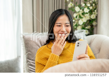 A cheerful woman is enjoying watching videos on social media on her smartphone on a sofa at home. A cheerful woman is enjoying watching videos on social media on her smartphone on a sofa at home. 116551257