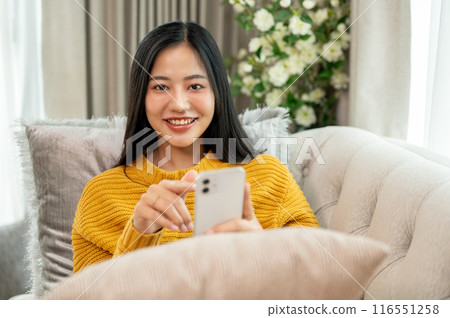 An attractive Asian woman is relaxing on a couch, holding her smartphone and smiling at the camera. An attractive Asian woman is relaxing on a couch, holding her smartphone and smiling at the camera. 116551258