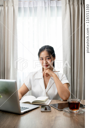 An attractive Asian businesswoman sits at her desk in her private office, holding a pencil. 116551380