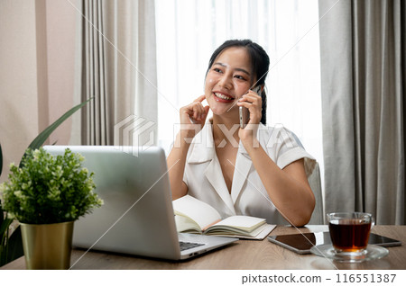 A confident Asian businesswoman is talking on the phone at her desk in her private office. 116551387