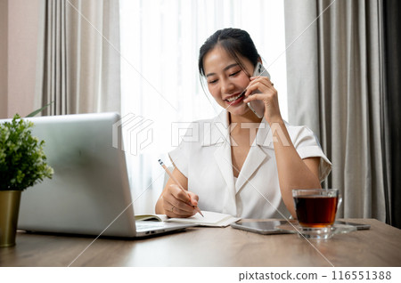 A beautiful Asian businesswoman is talking on the phone with her client while working at her desk. 116551388