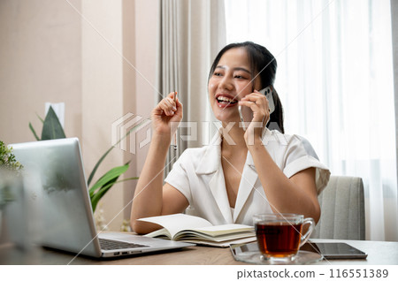 A confident Asian businesswoman is talking on the phone at her desk in her private office. 116551389
