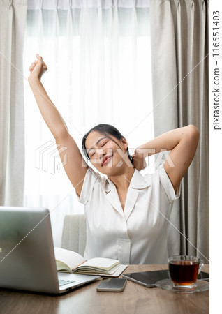 A carefree, happy young Asian woman is stretching her arms at her desk after finishing her work. 116551403