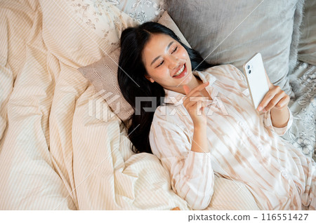 A beautiful Asian woman in pajamas chatting with her friends on her smartphone while lying in bed. A beautiful Asian woman in pajamas chatting with her friends on her smartphone while lying in bed. 116551427