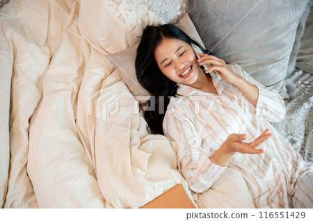 A charming Asian woman in pajamas is talking on the phone with her friends while lying in bed. 116551429