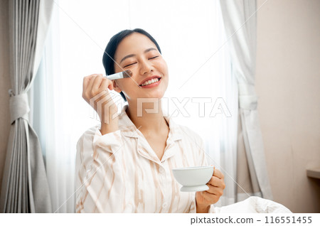 A beautiful, smiling young Asian woman in pajamas is applying a facial treatment mask to her face. 116551455