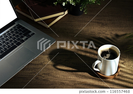 A cup of coffee laptop and books on wooden table lit by sunlight 116557468