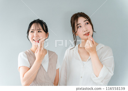 Woman putting on makeup and getting ready with her friend. Makeup image (lipstick, lip gloss, cosmetics, smiling asian woman Woman putting on makeup and getting ready with her friend. Makeup image (lipstick, lip gloss, cosmetics, smiling asian woman 116558139