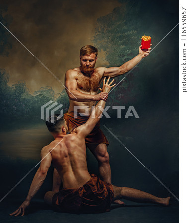 One man stands holding French fries high in air, while other kneels below, reaching up with longing expression against vintage background. 116559657