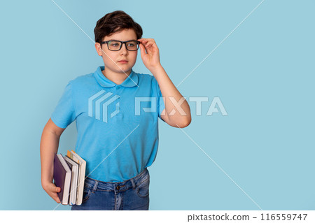 A cute teenage boy in glasses and a blue T-shirt, holding a stack of books 116559747
