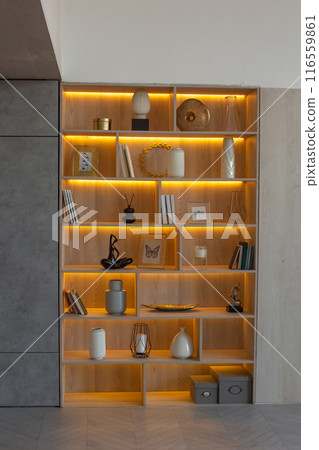 A shelving with figurines, vases and frames, with warm lighting, stands in a niche of the wall A shelving with figurines, vases and frames, with warm lighting, stands in a niche of the wall 116559861