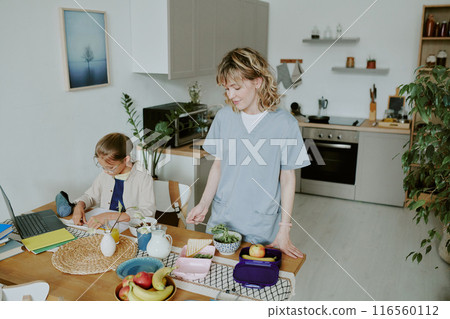 Young Caucasian doctor mom preparing two lunchboxes for her and her kid while her daughter doing homework Young Caucasian doctor mom preparing two lunchboxes for her and her kid while her daughter doing homework 116560112