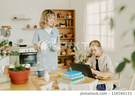 Good looking Caucasian nurse mom serving breakfast for her daughter while daughter making school project at dining table Good looking Caucasian nurse mom serving breakfast for her daughter while daughter making school project at dining table 116560133