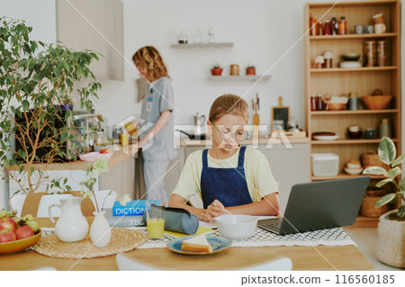 Caucasian girl sitting at table and doing homework while her doctor mother serving beverages before school and work Caucasian girl sitting at table and doing homework while her doctor mother serving beverages before school and work 116560185