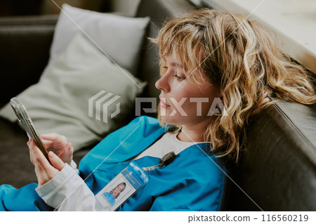 Medium close up of young blonde Caucasian nurse dressed in deep blue scrubs sitting on black leather sofa while checking her black smartphone Medium close up of young blonde Caucasian nurse dressed in deep blue scrubs sitting on black leather sofa while checking her black smartphone 116560219