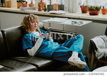 Focused blonde Caucasian nurse sitting on black sofa and surfing Internet while having small break during shift 116560382