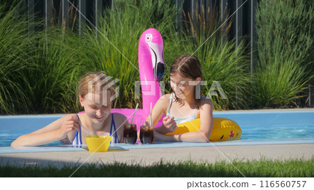 Two teenage girls in a pool with a pink flamingo float, enjoying beverages and snacks on a sunny day. 116560757