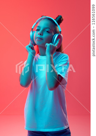 Portrait of young girl, child in casual clothes, white t-shirt listening to music in headphones against pink background in neon light 116561099