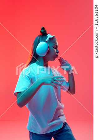 Portrait of young girl, child in casual clothes, t-shirt and jeans, listening to music in headphones and dancing against pink background in neon light Portrait of young girl, child in casual clothes, t-shirt and jeans, listening to music in headphones and dancing against pink background in neon light 116561104