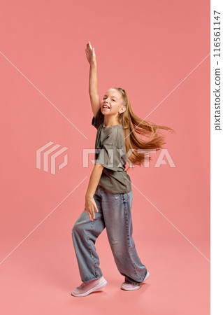 Full-length portrait of cheerful young girl, child in casual t-shirt and jeans, cheerful dancing and having fun against peach background 116561147