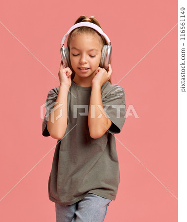 Portrait of young girl, child in casual clothes, t-shirt and jeans, listening to music in headphones against peach background Portrait of young girl, child in casual clothes, t-shirt and jeans, listening to music in headphones against peach background 116561149