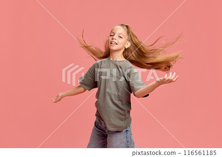 Portrait of young girl with long flowing hair, wearing grey t-shirt and jeans, posing against pink background. Joyful expression and happiness 116561185