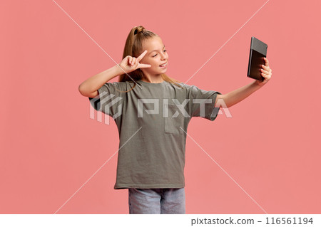 Portrait of young girl in casual clothes taking selfie with tablet, posing on social media, communicating and having fun against peach background Portrait of young girl in casual clothes taking selfie with tablet, posing on social media, communicating and having fun against peach background 116561194