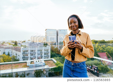 African American woman using smartphone, possibly checking her messages or browsing the internet on terrace. Female wearing a yellow jeans jacket and blue skirt. City skyline in background is visible 116563893
