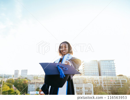 African female student graduate dressed in graduation cap and gown extends her hat to the camera and smiles. Study, education, university, college, graduate concept. 116563953
