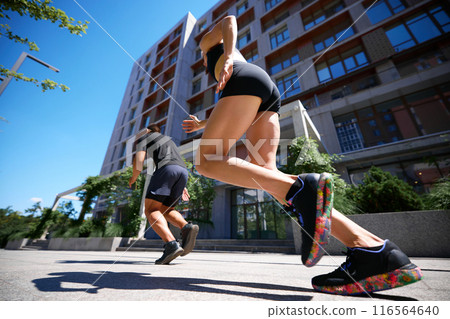 Low-angle view captures two runners sprinting in urban setting, with modern buildings towering above them. Fitness. Low-angle view captures two runners sprinting in urban setting, with modern buildings towering above them. Fitness. 116564640
