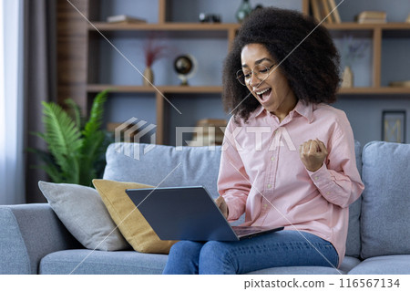 Happy woman with curly hair sits on sofa using laptop. Feeling excitement, happiness, success. Modern home environment with laptop, cushions, plants, shelves in background. 116567134